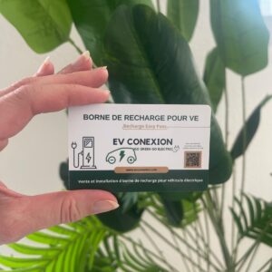 Hand holding a white EV charging card with 'Borne de recharge pour VE' and 'EV CONEXION' logo, charger icons, QR code, against a backdrop of green plants.