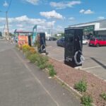 Parking lot with multiple electric vehicle charging stations in the foreground and a Feu Vert store in the background on a sunny day.