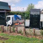 Electric vehicle charging kiosks (large black units) behind a green fence, with a white truck and blue lifting equipment in a construction‑like site.