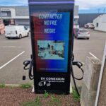 Electric vehicle charging kiosk in a parking lot; screen shows 'CONTACTER NOTRE REGIE' with an image of people at work and the EV CONEXION logo below.