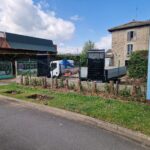 Construction area behind a green fence; white truck with a blue crane parked nearby against a stone building in the background.